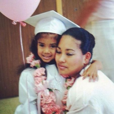 Mila J is wearing a graduation dress with a flower garland and hugging Christina Yamamoto.